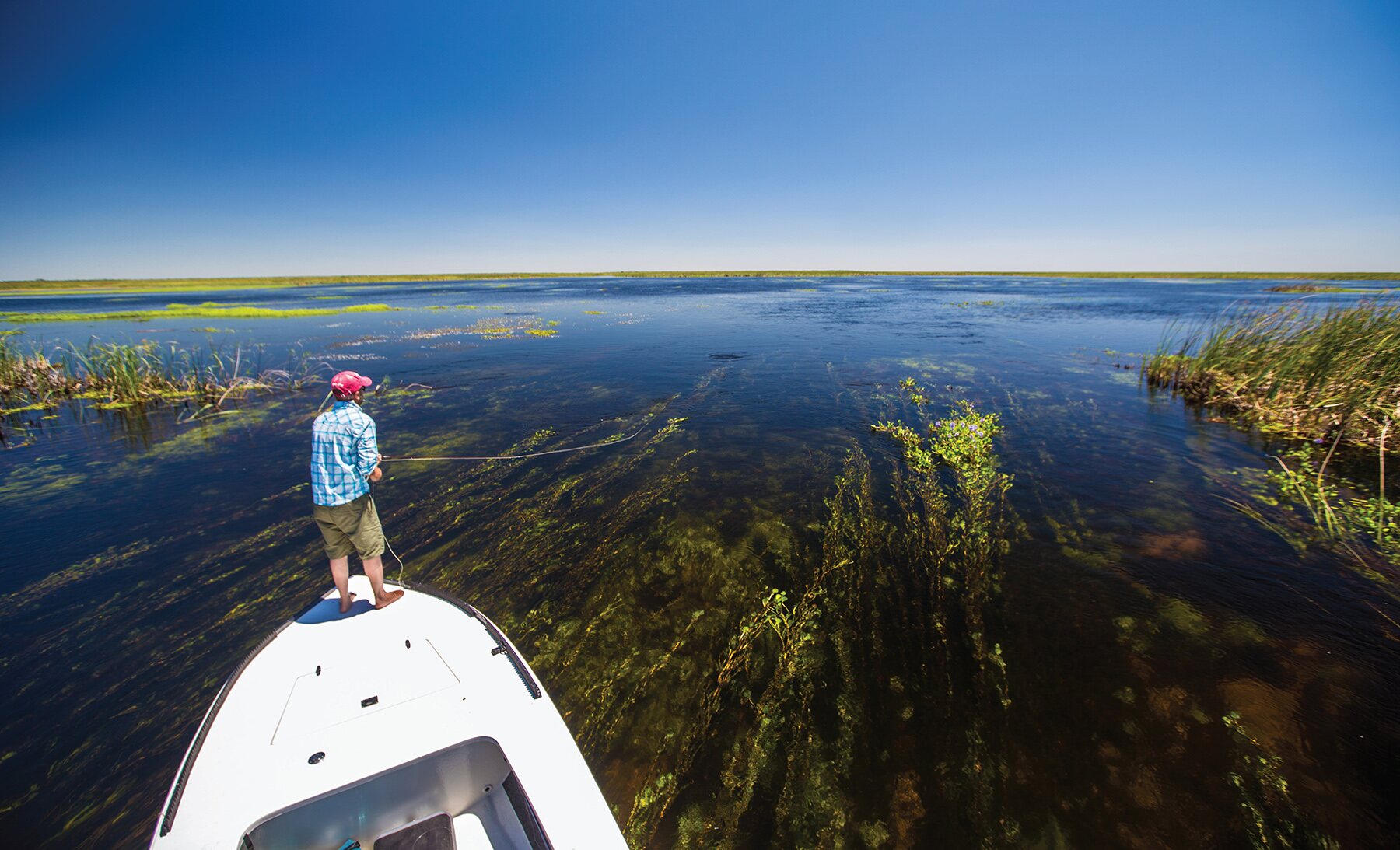 An angler on the bow of a skiff casting across still marsh flats